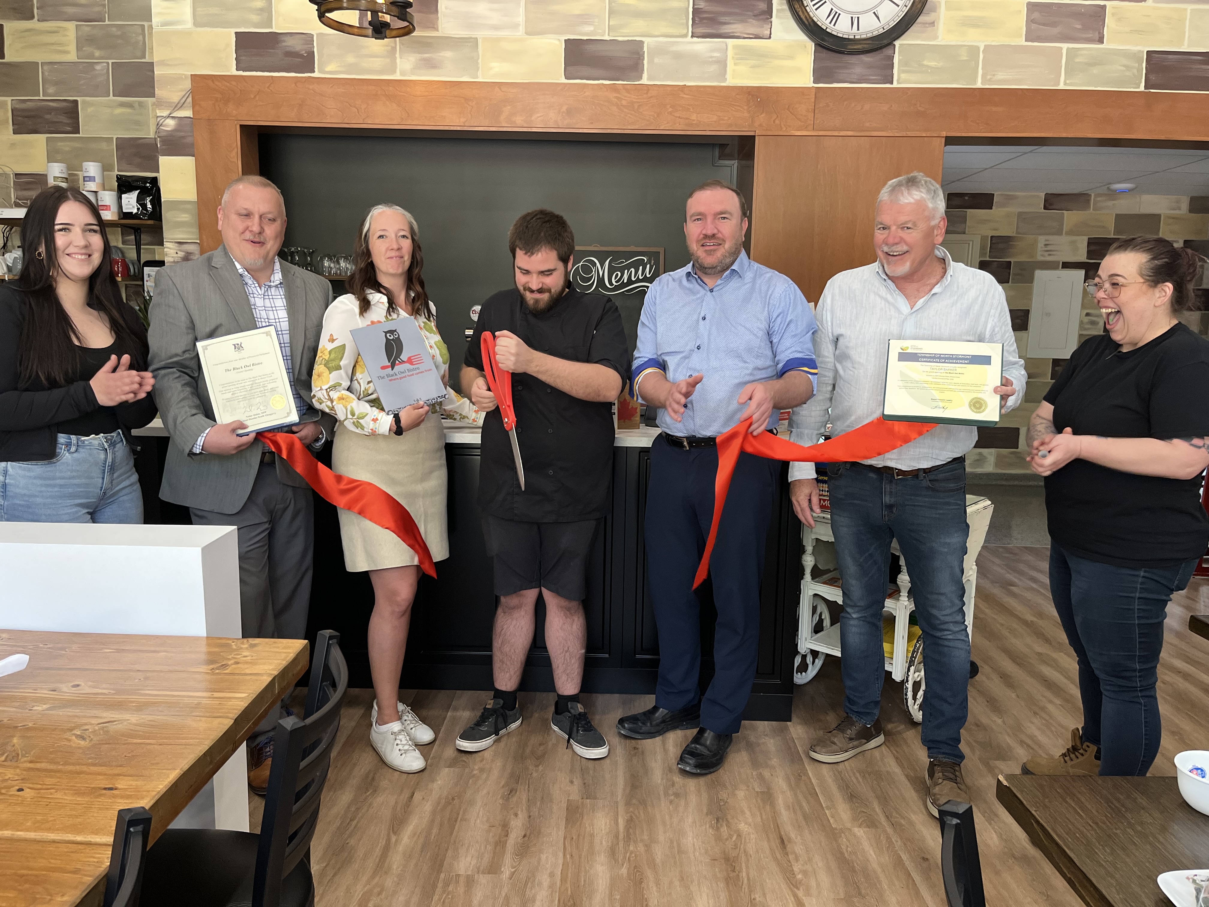 An image showing a group of people cutting a red ribbon to mark the opening of The Black Owl Bistro